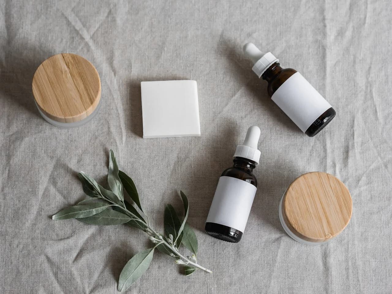 Produits cosmétiques naturels posés sur un tissu en lin — sérums en flacon ambré, savon blanc et boîtes en bambou avec une branche d'olivier